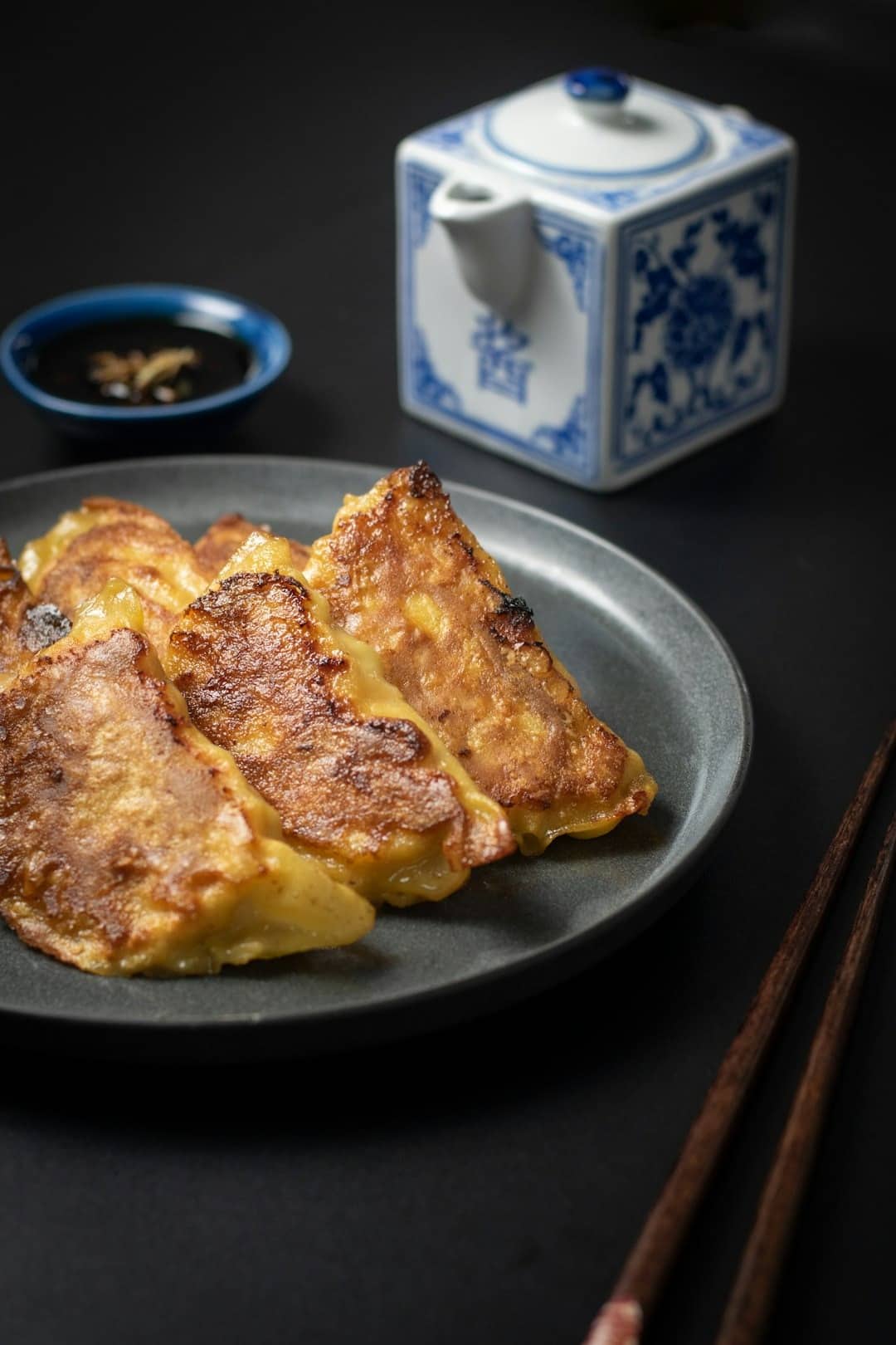 Golden-brown pan-fried dumplings on a dark plate with dipping sauce and chopsticks.